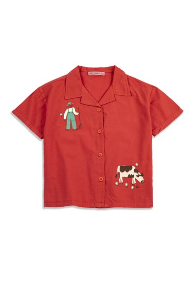 Chemise rouge à manches courtes avec col et boutons, ornée d'une illustration d'un fermier tenant une fourche et d'une vache en noir et blanc pâturant.
