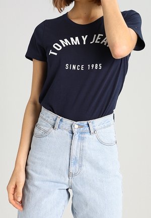 Tummansininen puuvillapaita, jossa on valkoinen "TOMMY JEANS" -teksti ja "SINCE 1985" -alateksti, yhdistettynä vaaleansiniin korkeavyötäröisiin farkkuihin.
