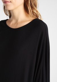 Svart, löst sittande topp med vid halsringning och draperade ärmar, tillverkad av mjukt tyg med en slät textur utan mönster.