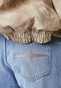 Poche de jean en denim bleu clair avec une broderie étoile à quatre branches centrée en rose, associée à un tissu beige froncé à la taille au-dessus.