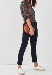Black and gold patterned blouse, navy trousers, and white espadrille sneakers. Brown crossbody bag with a curved design and yellow accent.