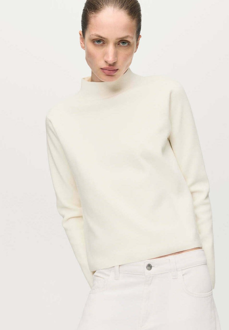 Femme à la peau claire et aux cheveux tirés en arrière, portant un pull à col roulé crème et un pantalon blanc sur un fond clair uni.