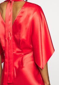 Robe en satin rouge avec un dos en V, nouée au cou avec un ruban. Présente des manches courtes et fluides ainsi qu'une texture lisse.
