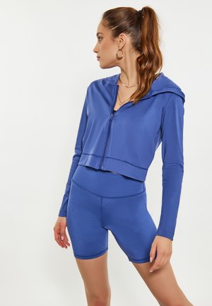 Kvinde med brunt hestehale iført blå cropped hættejakke med lynlås og matchende højtaljede cykelshorts, kigger mod venstre mod hvid baggrund.