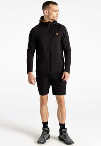 Schwarze Zip-Jacke mit orangen Akzenten, hohem Kragen und Seitentaschen, kombiniert mit schwarzen Shorts und grauen Wanderschuhen. Glatte Textur.