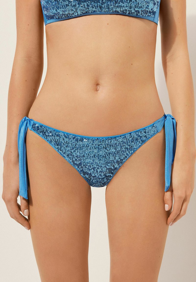 Blaue Bikiniunterteile aus einem strukturierten, schimmernden Stoff. Bänder an den Seiten mit einer glatten Oberfläche. Enthält eine Mischung aus hellen und dunklen Blautönen.