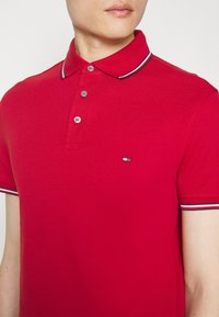 Personne portant un polo rouge avec des bordures rayées bleu marine, blanc et rouge au col et aux manches, arborant un petit logo rectangulaire sur la poitrine.