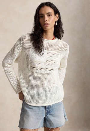 Γυναίκα με μακριά σκούρα μαλλιά που φοράει λευκό πουλόβερ ανοιχτής πλέξης και ανοιχτό μπλε τζιν σορτς, στέκεται μπροστά σε ουδέτερο φόντο.