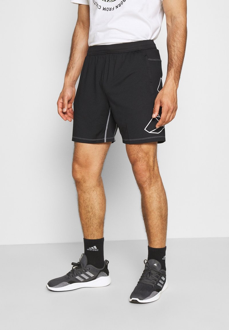 Shorts de sport noirs en tissu lisse, avec une taille élastique et des coutures blanches contrastantes ainsi que des accents de design géométrique. Portés avec des baskets sombres.