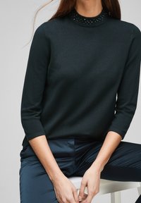 Pull vert foncé avec un col montant, orné d'une bordure cloutée. Fabriqué en tissu tricoté à la texture lisse avec des manches trois quarts.