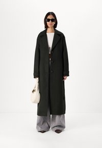 Μακρύ, σκούρο πράσινο παλτό με μεγάλα καπάκια και κλείσιμο με κουμπιά. Συνδυάζεται με γκρίζες φαρδιές παντελόνες και μαύρες μυτερές μπότες. Κρατάει μια κρέμα τσάντα.