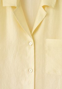Chemise jaune clair avec un col, deux boutons blancs et une poche poitrine, présentée à plat avec de légers plis de tissu.