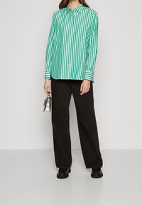 Camicia button-up a righe verdi e bianche con maniche lunghe, indossata con pantaloni neri a gamba larga e che tiene una piccola borsa a tracolla argentata.