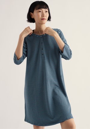 Femme aux cheveux courts noirs portant une robe rayée bleue et noire longueur genoux avec des boutons, tenant l'encolure avec les deux mains.