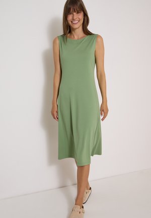 Kvinde iført ærmeløs lys grøn midi-kjole og beige slip-on sko står mod en ensfarvet lys væg.