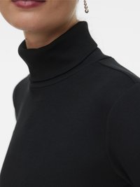 Közelkép egy fekete bordázott garbót viselő személyről, aki lógó gyöngy fülbevalót visel, az ajka és az alsó arca részben látható.