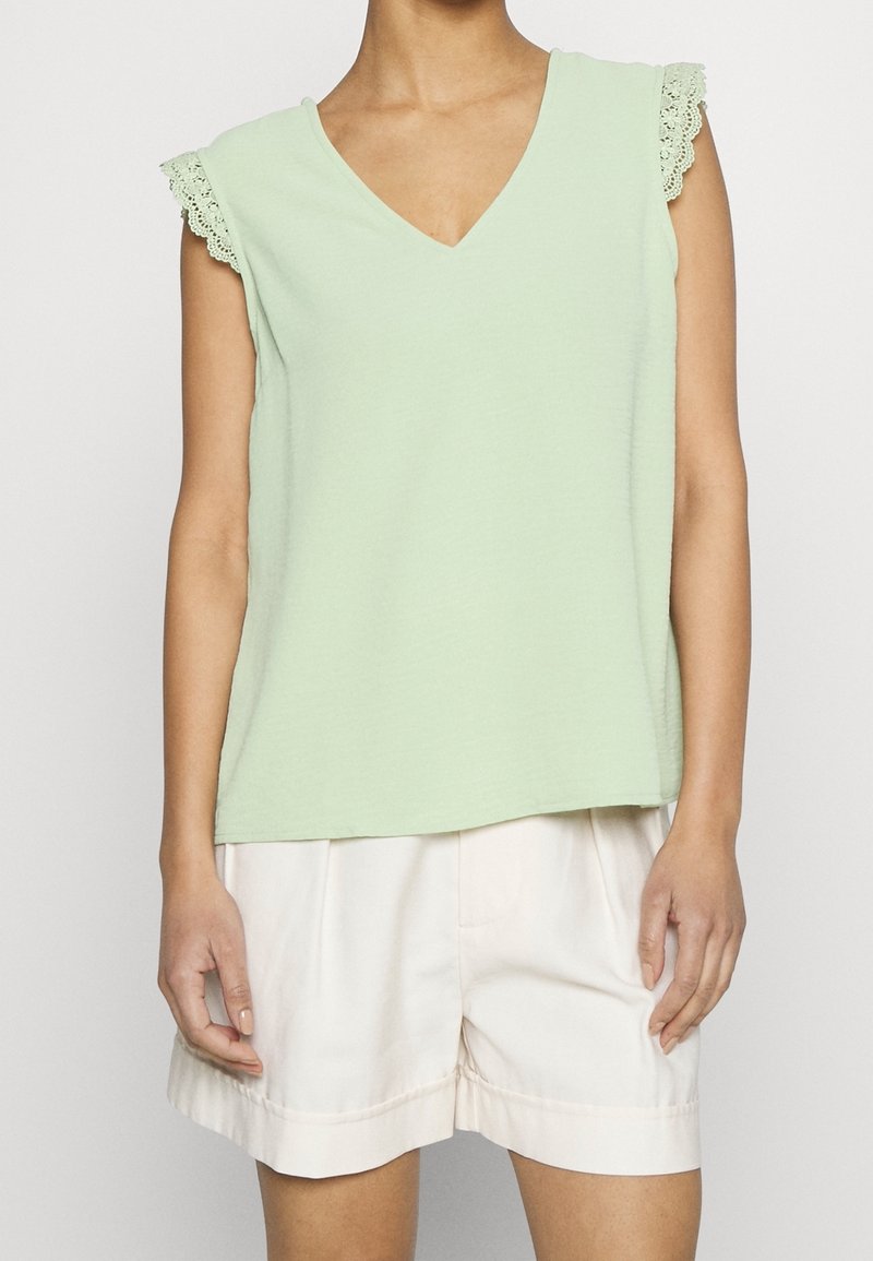 Haut sans manches vert clair avec un col en V et des détails en dentelle sur les épaules. Tissu doux avec une coupe décontractée, associé à un short crème clair.