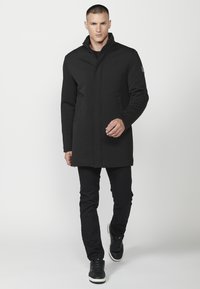 Cappotto lungo nero con colletto alto, design strutturato e due tasche laterali, abbinato a pantaloni neri e sneakers scuri.