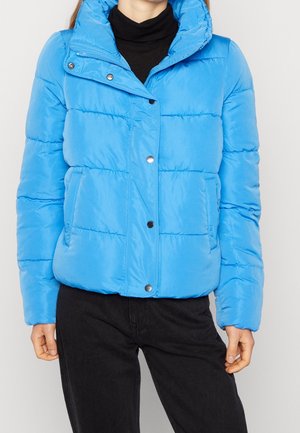 Personne portant une doudoune bleu vif avec des boutons-pression sur un col roulé noir et un pantalon noir, debout devant un fond uni.