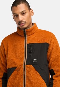 Mann trägt eine orange Fleecejacke mit schwarzer Brusttasche und Timberland-Logo, blickt nach vorne mit neutralem Ausdruck.