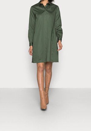 Langärmliges, dunkelgrünes Kleid mit Kragen, vorn mit Knopfleiste und seitlichen Taschen. Kombiniert mit beigefarbenen Stiefeletten.