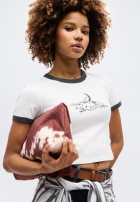 Νεαρή γυναίκα με σγουρά μαλλιά, που φοράει λευκό crop top Levi’s και κρατάει ένα καφέ μπουφάν με σχέδια, μπροστά σε απλό φόντο.