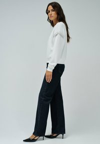 Sweat-shirt blanc avec des accents en strass sur les manches, associé à un jean noir à jambes larges et des talons pointus noirs. Texture lisse et design décontracté.