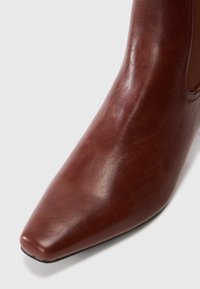 Acercamiento de una bota de cuero marrón hasta el tobillo con punta cuadrada y panel lateral elástico sobre un fondo blanco.