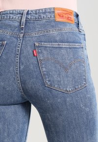 Jeans de mezclilla azul con un diseño ajustado, que presentan una etiqueta de cuero, bolsillos traseros con un patrón de costura curvada y una pequeña etiqueta de logo roja.