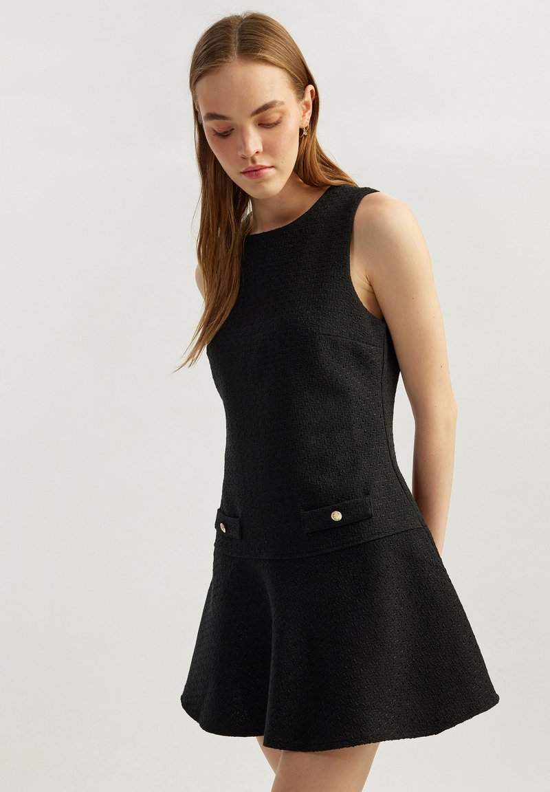 Vestido negro hecho de tela texturizada, con diseño sin mangas, falda acampanada, dos bolsillos frontales con detalles en botones dorados.