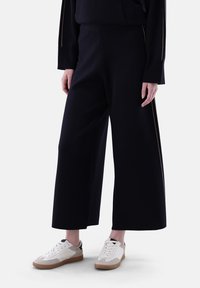 Schwarze weit geschnittene Hosen aus einem glatten Stoff, mit einem goldenen Akzent entlang der Seite. Kombiniert mit weißen und beigen Sneakern.