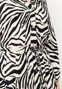 Blusa a righe zebrate in bianco e nero con colletto, chiusura a bottoni e vita elastica con cintura legata. Tessuto leggero e liscio.