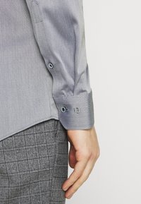 Camicia grigia chiaro in tessuto intrecciato con maniche lunghe, polsini con bottoni e tessuto strutturato, abbinata a pantaloni grigi a quadri.