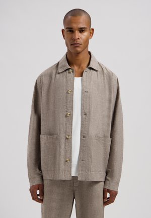 Man draagt een beige gestreept jasje met knopen over een wit shirt, gecombineerd met bijpassende beige gestreepte broek, staand tegen een effen achtergrond.