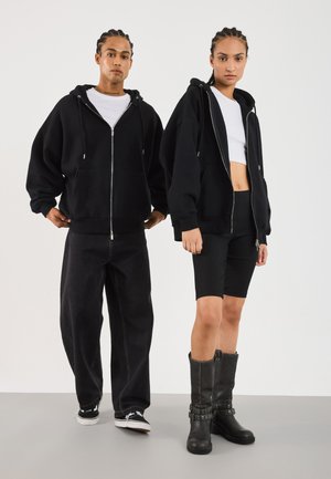 Sudadera negra con cremallera y corte oversize, combinada con jeans negros y un top corto. El modelo a la derecha lleva pantalones cortos de canalé y botas altas con tachuelas.