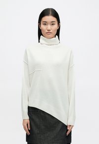 Λευκό πουλόβερ με λαιμόκοψη τύπου turtleneck, με διαγώνια ραφή, πλέξη και ριγέ τελείωμα. Φοριέται πάνω από μια σκούρα γκρι φούστα.