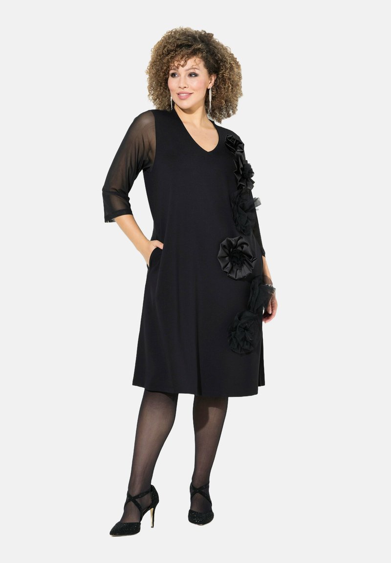 Schwarzes Kleid mit V-Ausschnitt und dreiviertellangen, durchsichtigen Ärmeln. Verfügt über Stoffrosen auf der linken Seite und seitliche Taschen. Trägt schwarze High Heels.