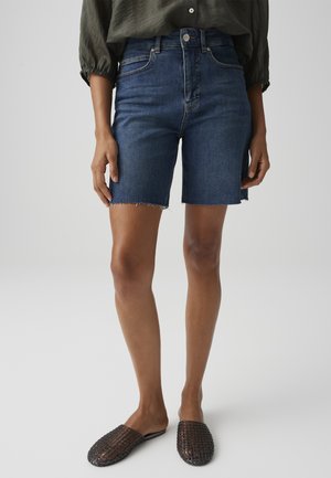 Persona che indossa pantaloncini di jeans blu scuro al ginocchio con orlo grezzo e scarpe slip-on intrecciate scure su sfondo semplice.