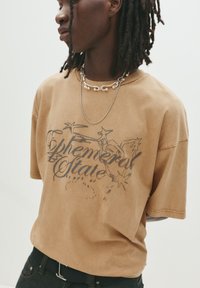 Brun bomulds t-shirt med korte ærmer, der har et sort grafisk print med teksten "Ephemeral State" og en afslappet pasform. Accentueret med et sølvfarvet kædethalskæde.