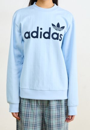 Osoba ubrana w jasnoniebieską bluzę Adidas z czarnym logo i luźne spodnie w kratę, stojąca na tle jednolitym.