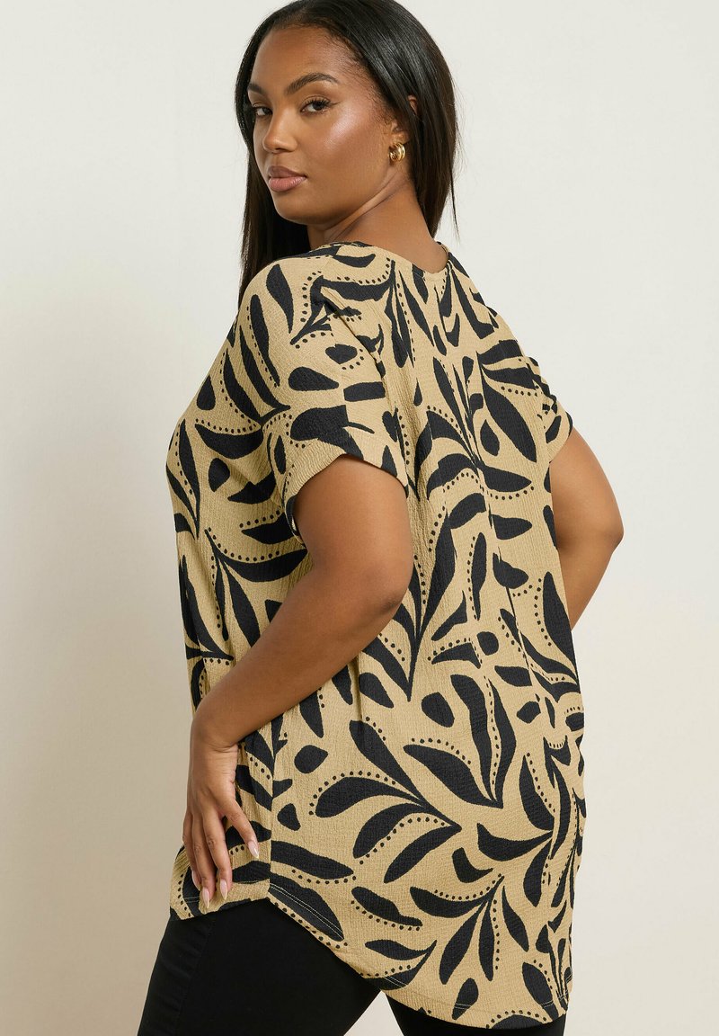 Mujer con cabello largo, vistiendo una camisa beige con patrones abstractos de hojas negras, posando con una mano en la cadera y mirando por encima del hombro.