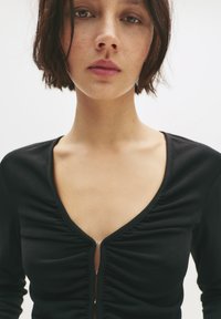 Top nero aderente con maniche lunghe, scollatura a V profonda, dettagli arricciati e zip frontale. Tessuto liscio, aderente al corpo, design minimal.