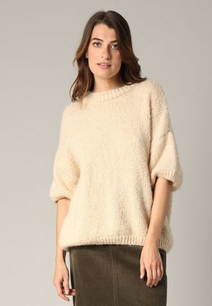 Pull en maille flou couleur crème, avec un col rond et des manches courtes, associé à une jupe en velours côtelé de couleur vert olive. Texture douce.