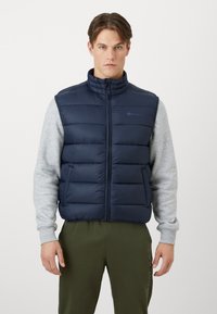 Navyfarbene Steppweste mit hohem Kragen, gestepptem Muster und Reißverschluss auf der Vorderseite. Getragen über einem hellgrauen Langarmshirt mit gerippten Bündchen.