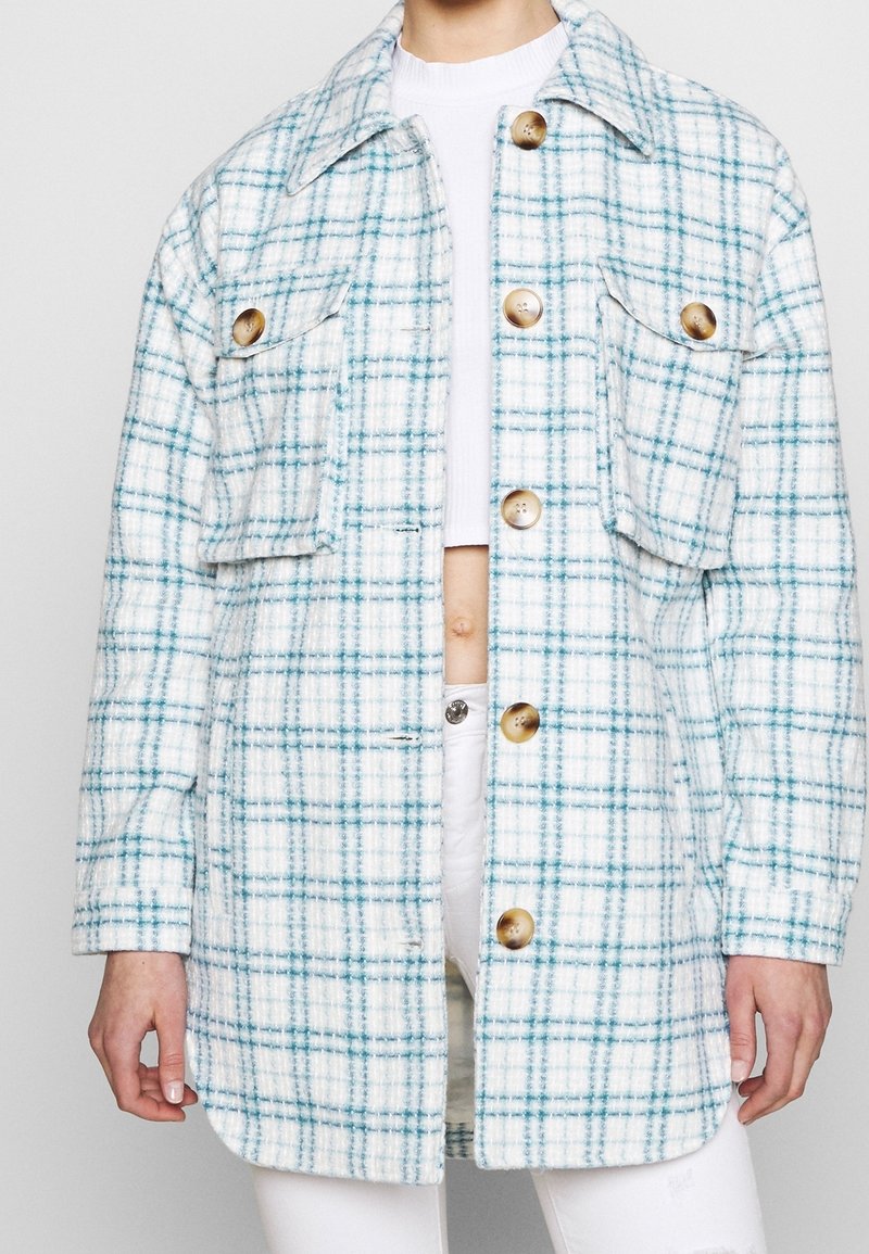 Koszula-ramoneska w kratę o białej bazie z niebieskimi liniami, dwie kieszenie na piersi, duże guziki i luźny krój. Wykonana z teksturowanej tkaniny.