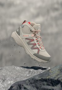 Ανοιχτό γκρι και κόκκινο ορειβατικό μποτάκι με παχιά σόλα, αιωρούμενο στον αέρα πάνω από βραχώδη έδαφος, με κόκκους βρωμιάς να πέφτουν κάτω.