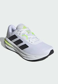 Zapatilla deportiva blanca con rayas negras, detalles en verde neón, parte superior de malla, suela texturizada y collar acolchado para mayor comodidad.