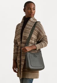 Borsa a mano in pelle grigia con forma strutturata, logo in rilievo e tracolla regolabile. Il modello indossa un cardigan beige a fantasia e un top scuro.