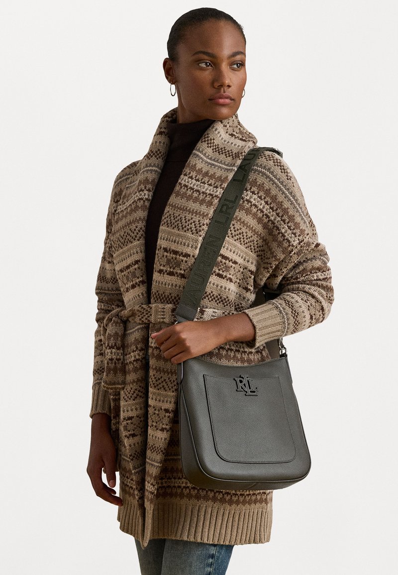 Borsa a mano in pelle grigia con forma strutturata, logo in rilievo e tracolla regolabile. Il modello indossa un cardigan beige a fantasia e un top scuro.