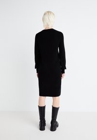 Knit black dress with long sleeves and fitted design, knee-length. Worn with tall black boots featuring buckles. Smooth texture, minimal detailing.